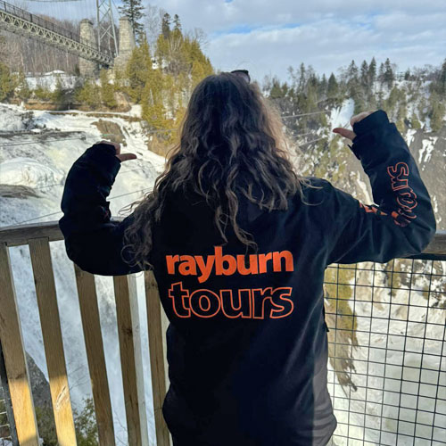 Rayburn Tours Ski Rep Annabelle Nojac poses for the camera, with her face towards Montmorency falls, and her back, with the Rayburn Tours orange logo on the back of her ski jacket, towards the camera