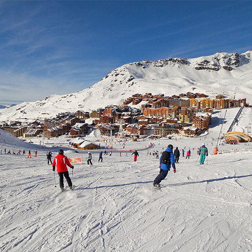 Skiers ski down a slope towards a resort in the background.