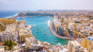 An aerial view of Spinola Bay in St. Julians, Malta. The bay is filled with boats, and the surrounding area is densely populated with colorful buildings.