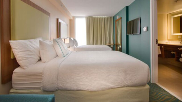 A hotel room with two double beds, a television, a window with curtains, and a door leading to a bathroom. The walls are painted white, and the headboard is a teal color.