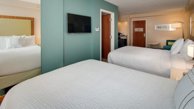 A hotel room with two double beds, a television, a door leading to a bathroom and kitchenette area, and a window with curtains. The walls are painted teal, and the headboard is a light brown color.