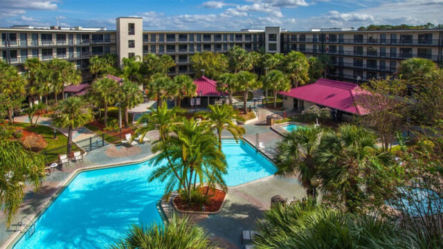 An aerial view of a hotel complex with multiple buildings surrounding a large, irregularly shaped pool. Palm trees and other tropical plants are scattered throughout the property.