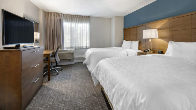 A hotel room with two double beds, a desk, a dresser, a television, and a window with curtains. The walls are painted white, and the headboard is a dark wood color.