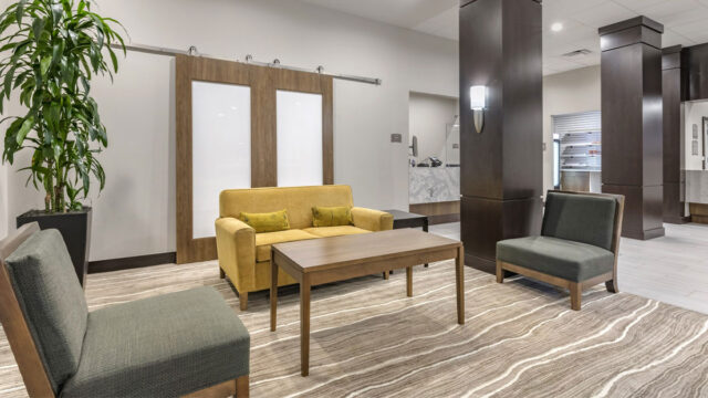 A hotel lobby with a seating area featuring a yellow couch, armchairs, and a coffee table. There are tall columns and a sliding glass door in the background.